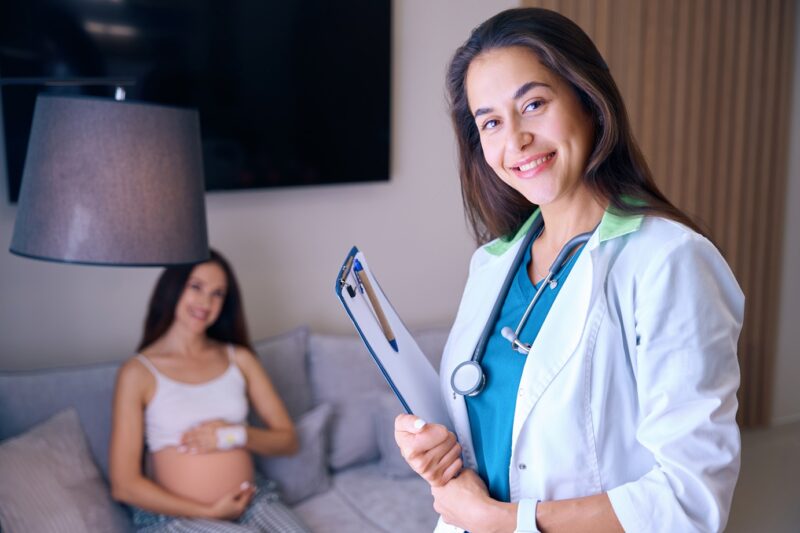 female nurse with clipboard looking at camera near blurred pregnant woman